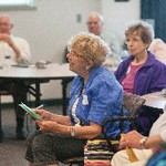 A seated woman holding papers is intently looking up, while others are seated at a table next to her